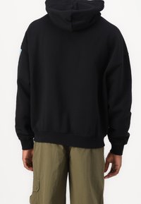 Schwarzer Baumwoll-Hoodie mit lockerem Schnitt, ausgestattet mit einer Kängurutasche und gerippten Bündchen. Rückansicht zeigt ein nahtloses Design und Kapuze.