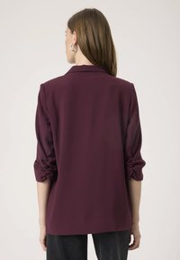 Burgundia bleiser, mille üleskeeratud varrukad, kohandatud lõige ja sile kangas. Omab nooled lapelit ja puhta, lihtsa selja disaini.