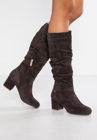 Mujer vestida con botas de ante marrón oscuro hasta la mitad de la pantorrilla, con tacones de bloque y cremalleras laterales, de pie sobre un suelo claro frente a una pared blanca lisa.