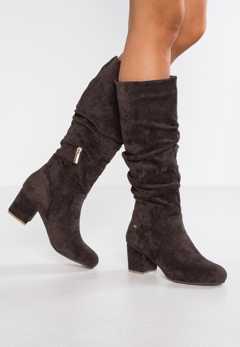 Mujer vestida con botas de ante marrón oscuro hasta la mitad de la pantorrilla, con tacones de bloque y cremalleras laterales, de pie sobre un suelo claro frente a una pared blanca lisa.