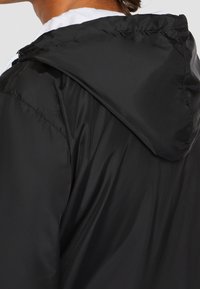 Zwarte capuchonjack met een gladde, glanzende textuur en een ontspannen pasvorm. Gedetailleerde stiksels op de schouders en rondom de capuchon.