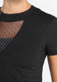 Svart T-shirt med rund halsringning, texturerad meshpanel med prickar och en diamantmönstrad infällning vid bröstområdet.
