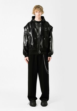 Sudadera con acolchado negro y acabado brillante, de corte oversize y bolsillos con cremallera. Combinada con pantalones negros de corte holgado y zapatos negros gruesos.