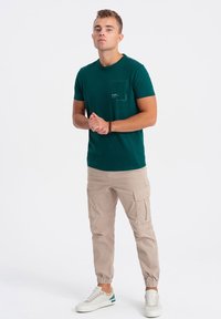 Tyrkysové tričko s kulatým výstřihem a kapsou, v kombinaci s béžovými cargo kalhotami s elastickými manžetami a bočními kapsami, a bílými teniskami.