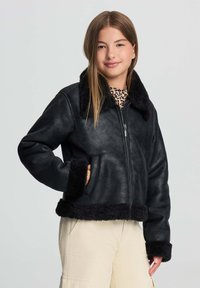 Giovane che indossa una giacca in shearling nera, una maglietta con stampa leopardata e pantaloni beige, in piedi con una mano in tasca su uno sfondo chiaro.