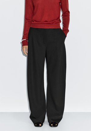 Un pull rouge à manches longues, associé à un pantalon noir à jambes larges. Le pantalon présente des poches latérales et une texture de tissu lisse.