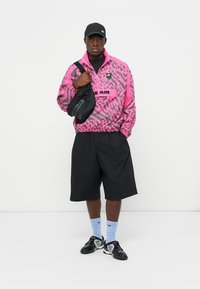 Pink patterned windbreaker with black accents, black shorts, and black sneakers. The person also carries a small black crossbody bag.