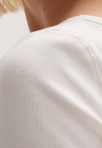 Vitt ribbat stickat plagg med rund halsringning, som visar en närbild av ärmen och axeldetaljen, vilket framhäver den fina texturen.