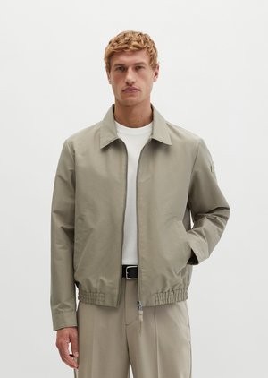 Uomo che indossa giacca beige con zip sopra camicia bianca e pantaloni beige abbinati, in piedi davanti a uno sfondo chiaro e uniforme.