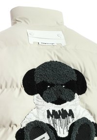 Chaqueta acolchada en color crema con un perro de dibujos animados en relieve en negro y blanco bordado en la parte posterior, que presenta un cuello blanco y un detalle de marca.