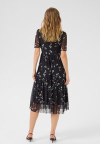 Rochie neagră florală cu mâneci scurte transparente, fustă cu volane în straturi și talie ajustată. Prezintă modele florale mici în alb și roșu.
