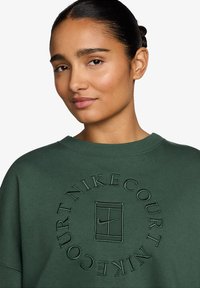 Sudadera de algodón verde con un gráfico redondo en relieve que presenta el texto y logo "Nike Court" en el centro. Cuello redondo acanalado y puños.