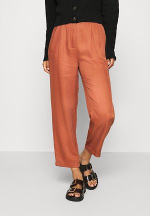 Person trägt hoch taillierte rostfarbene Hose, schwarzen, geknöpften Pullover und schwarze Sandalen mit offenen Zehen und zwei Schnallenriemen.