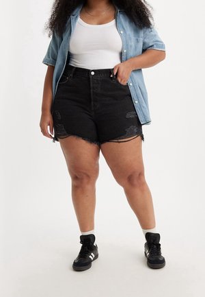 Kvinde iført sorte slidte denimshorts, hvid tanktop, lyseblå kortærmet skjorte og sorte sneakers med hvide striber.