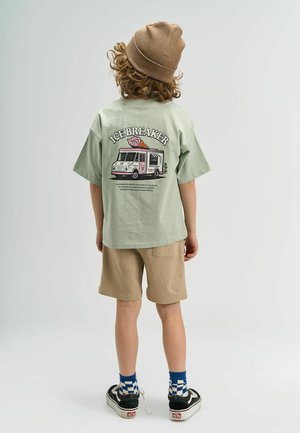 Göndör hajú gyerek, barna sapkát visel, zöld "Ice Breaker" póló van rajta, amelyen egy fagylaltos kocsi grafika látható, bézs rövidnadrág, kockás zoknik és fekete sportcipő.