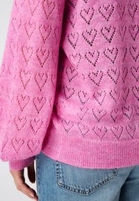 Roze gebreide trui met hartpatronen, met een textuur en open breiwerk ontwerp en geribbelde mouwen, gedragen over blauwe denim jeans.