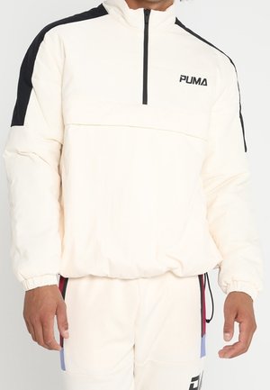 Hombre vestido con un pulóver blanco de Puma con rayas negras en los hombros y una cremallera frontal, combinado con pantalones blancos a juego.