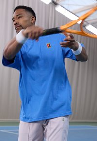 Lyseblå t-skjorte med kort erme og en liten flerfarget logo på brystet, sammen med lysegrå shorts, holder en tenniskølle.