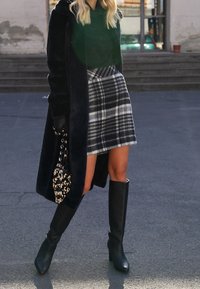 Langer schwarzer Kunstpelzmantel, grüner Pullover, karierten Minirock in Schwarz und Weiß, knielange schwarze Stiefel mit Absatz und eine gemusterte Handtasche.