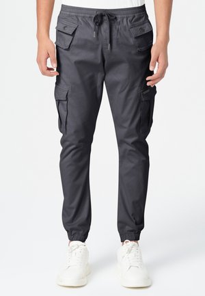 Pantalons cargo gris foncé avec des poignets élastiques, des poches latérales et une taille à cordon, fabriqués en tissu léger. Baskets blanches en dessous.