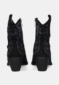 Par de botines de ante negro con tacones de bloque medianos, cremalleras laterales y studs decorativos negros en patrones intrincados.