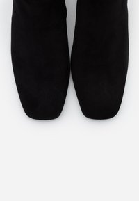 Bottes chevilles noires en daim avec une texture lisse, présentant un design à bout carré et une surface unie sans ornements supplémentaires.