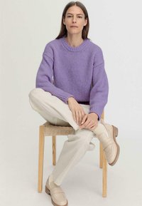 Laventeli neulepaita pyöreällä pääntiellä, yhdistettynä kermavalkoisiin leveälahkeisiin housuihin ja beigenvärisiin loafereihin, istuu puutuolilla.