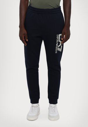 Muž v černých upravených teplácích s bílým logem Emporio Armani na stehně a bílých teniskách, stojící před jednobarevným pozadím.