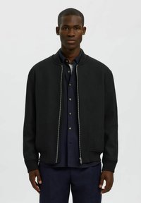 Giubbotto bomber nero con zip frontale, polsini a coste e colletto tondo, realizzato in tessuto testurizzato, indossato sopra una camicia blu scuro.