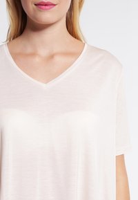 Kvinna med ljus hy och blont hår som bär en ljus rosa t-shirt med V-ringning och korta ärmar mot en enkel vit bakgrund.