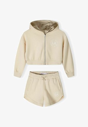Felpa beige in cotone misto con zip frontale e logo, abbinata a pantaloni corti coordinati con vita elastica e tasche laterali.