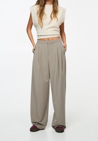 Lichtbeige mouwloze gebreide top gecombineerd met hoge taille, wijde taupe broek. De broek heeft voorvouwen en zijzakken.