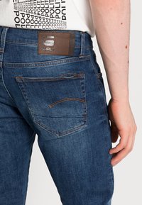 Jeans de mezclilla en azul oscuro, con costuras naranjas, una parche de cuero marrón y un diseño ajustado con bolsillos traseros discretos.