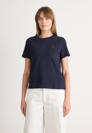 Mörkblå bomulls-t-shirt med korta ärmar, rund halsringning och en liten röd logotyp på bröstet, i kombination med vita vida byxor.