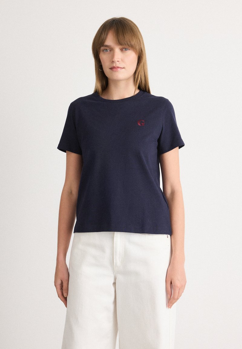 T-shirt en coton bleu marine à manches courtes, col rond, avec un petit logo rouge sur la poitrine, associé à un pantalon large blanc.