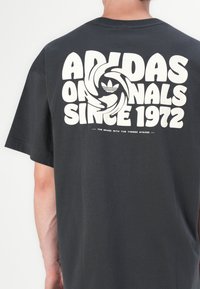 Tamno siva majica s velikim bijelim grafičkim natpisom na leđima, uključujući "Adidas Originals Since 1972" i Adidas logotip.