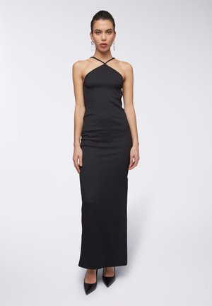 Abito lungo senza maniche di colore nero con una silhouette aderente, tessuto testurizzato e spalline incrociate. Indossato con tacchi neri a punta e orecchini a forma di stella.
