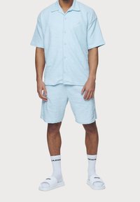 Vaaleansininen teksturoitu lyhythihainen paita ja siihen sopivat shortsit. Yksityiskohtina napit, pieni logo, yhdistetty valkoisiin liukuisiin sandaaleihin ja sukkiin.