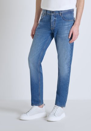 Jeans en denim bleu clair avec une coupe droite, présentant un design classique à cinq poches, une fermeture éclair et des détails de décoloration subtils, associés à des baskets blanches.