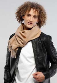 Beige Wollschal mit fransigen Enden, locker um den Hals gewickelt, kombiniert mit einer schwarzen Lederjacke und einem weißen Hemd. Lockiges Haar ist sichtbar.