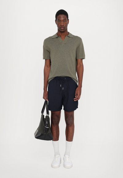 Camisa polo verde-azeitona, calções azul-marinho, meias brancas e ténis brancos. Os acessórios incluem uma mala verde-escura com uma base preta.