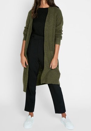 Žena v dlouhém olivově zeleném kardiganu, černém topu a kalhotách, s bílými nazouvacími teniskami, stojící před jednobarevným pozadím.