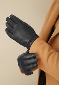 Guanti in pelle nera con una texture liscia, caratterizzati da un design tridimensionale e dita leggermente curve. Indossati con un cappotto color cammello.
