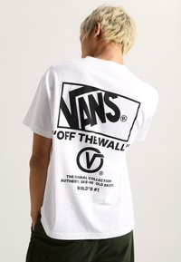 T-shirt en coton blanc avec un imprimé graphique noir "VANS" et "OFF THE WALL" au dos, avec des détails de texte et de logo en dessous. Manches courtes.