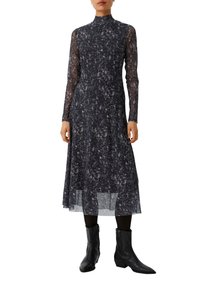 Schwarzes langärmliges Kleid mit hohem Kragen, das ein strukturiertes weißes Muster aufweist, einen geschichteten Rock hat und mit schwarzen Cowboys Stiefeln kombiniert ist.