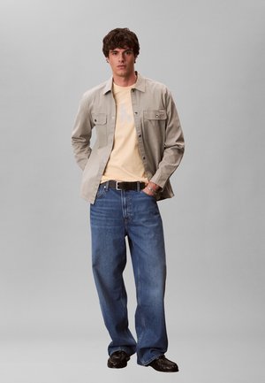 Jeune homme debout, mains dans les poches, portant une chemise beige boutonnée sur un t-shirt jaune, un jean bleu ample, une ceinture noire et des chaussures noires.