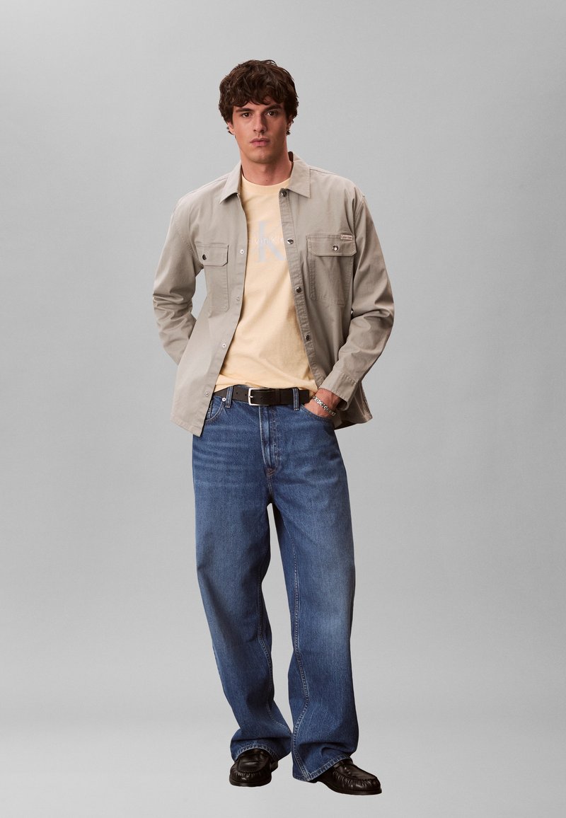 Noor mees seisab käed taskutes, seljas beež nööbitav särk kollase t-särgiga, jalas laiad sinised teksad, must vöö ja mustad kingad.