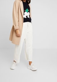 Donna in cappotto beige, maglione nero con design di orso polare bianco e albero di Natale, pantaloni bianchi a coste e sneakers bianche, in piedi su un pavimento grigio.