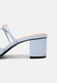 Světle modré sandály na blokovém podpatku se dvěma tenkými pásky, které mají malou růžovou květinovou ozdobu a černou podrážku. Hladká kožená textura.