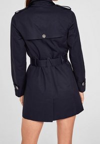 Ciemnoniebieski płaszcz trenchowy z paskiem w talii, z guzikiem, naramiennikami i wentylacją z tyłu dla swobody ruchów. Gładkie wykończenie tkaniny.
